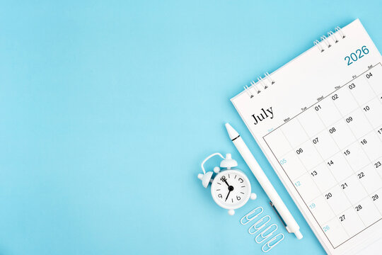 July 2026, Monthly desk calendar for 2026 year and alarm clock with paper clips on blue color background.