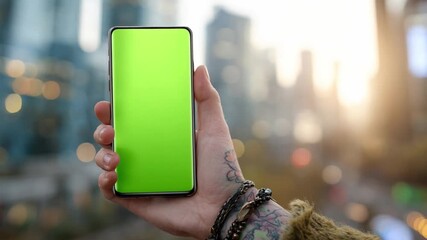 Smartphone with Green Screen: A hand cradles a sleek smartphone with a vibrant green screen, set against a blurred city skyline backdrop, ready for digital content. 