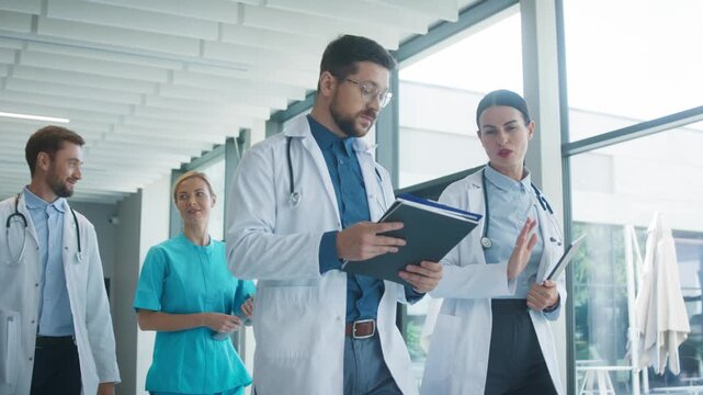 Medical staff walking through bright clinic hallway talking with each other. Focused male doctor reviewing notes while colleague discuss plan. Nurse joining conversation and sharing thoughts.
