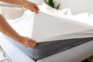 Changing bed sheets. Woman is putting on a fitted white cotton sheet on a mattress while making the...