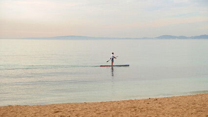 Man paddleboarding alone on calm sea. Soft pastel horizon. Concept of freedom, balance, mindfulness and healthy coastal lifestyle.