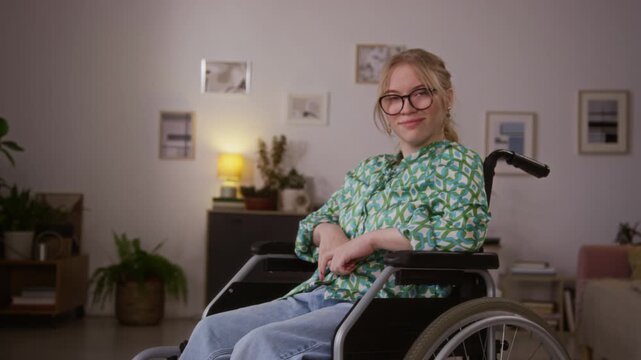 Portrait of Caucasian young woman with blonde hair with paraplegia looking at camera while sitting in wheelchair in her apartment