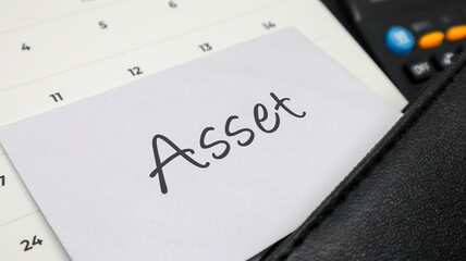 Handwritten word ASSET on a white note placed on a calendar with a calculator and leather planner.