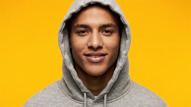 Young biracial man in a grey hoodie transitions from a serious expression to a happy smile, looking directly at the camera against a vibrant yellow background.