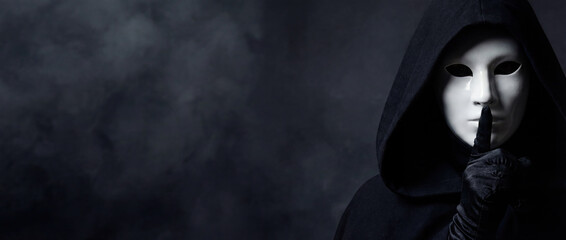 Mysterious figure in white Venetian mask and black hooded cloak making silence gesture. Anonymous person keeping a secret on dark smoky background. Masquerade and drama concept.