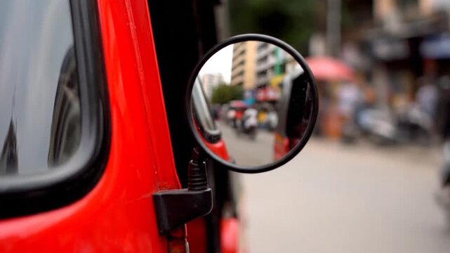Close-up of a red auto-rickshaw side mirror reflecting a bustling city street with traffic, motorcycles, and people. Captures urban life and movement.