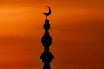 Silhouette of mosque crescent moon finial stands against vibrant orange sunset sky, creating serene and spiritual atmosphere. intricate design of finial is highlighted by contrasting colors