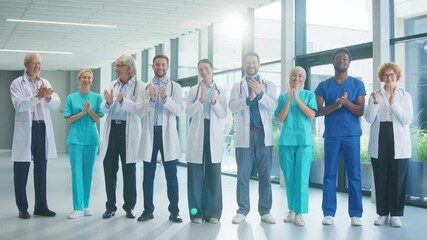 Medical team standing in bright hospital hall clapping toward camera. Smiling proudly. Showing appreciation for teamwork and celebrating achievements while enjoying positive moment.