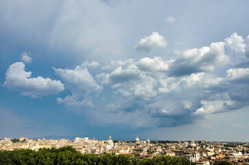 Naklejka premium Wide panoramic Rome skyline with Vittoriano monument, baroque church domes and terracotta rooftops under dramatic blue sky filled with billowing white and grey storm clouds