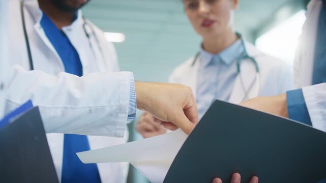 Close view of doctors exchanging papers in bright hospital hall. Reviewing lab findings. Pointing at data and clarifying details. Coordinating patient care while preparing next steps together.