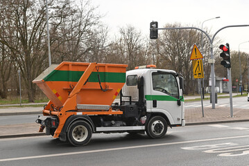 A white garbage truck with an orange skip container driving on a city road near a traffic light. Professional waste management vehicle in an urban environment.