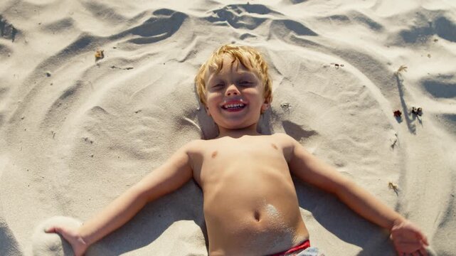 Smiling Little Boy In Swimsuit Lies On Warm Yellow Sand And Makes A Sand Angel By The Sea. Summer Beach Vacation Scene With Childhood Joy, Family Travel, And Carefree Outdoor Fun.