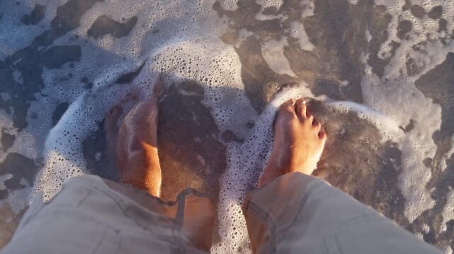 POV view shows bare feet on wet sand as crystal-clear water gently washes over ankles. A peaceful summer vacation moment with soft waves, slow steps, and oceanfront serenity.