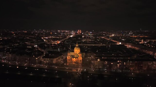Night drone view of Munich featuring the iconic twin towers of Frauenkirche on the horizon. St. Lukas Church stands by the Isar river, with the illuminated Old Town glowing in the distance