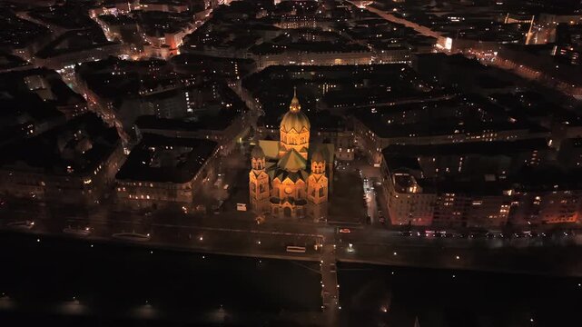 Naechtliche Luftaufnahme von Muenchen mit den markanten Tuermen der Frauenkirche am Horizont. Im Vordergrund die St. Lukas Kirche an der Isar und die beleuchtete Silhouette der Altstadt