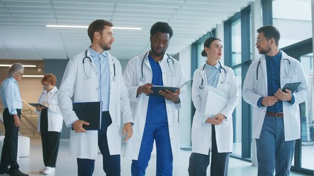 Group of doctors walking along bright hospital corridor. Holding tablet and folders. Talking and sharing updates. Planning tasks for shift and coordinating teamwork during busy morning.