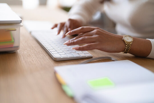 A person wearing a watch types on a keyboard, with a notebook and sticky notes on a wooden desk.