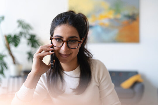 A woman with glasses smiles while talking on her mobile phone in a bright, modern room with plants and artwork.