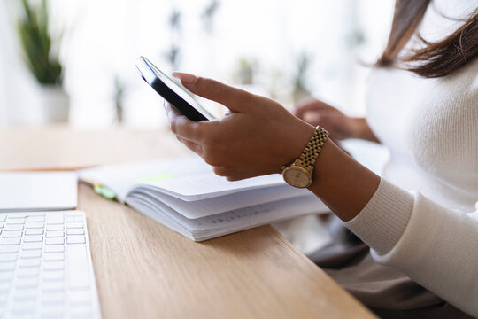 A person wearing a watch uses a smartphone while sitting at a desk with a notebook and keyboard.