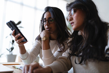 Two women collaborate on a project, one holding a smartphone while the other looks on thoughtfully, suggesting teamwork and shared ideas.