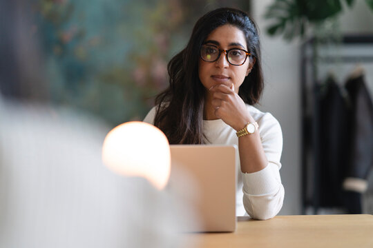 A thoughtful woman wearing glasses sits at a desk with a laptop, her hand on her chin, contemplating her work.