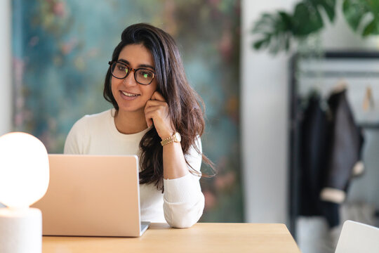 A young woman with glasses smiles while working on her laptop at a desk, with a lamp and artwork in the background.