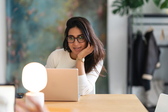 A woman wearing glasses smiles while looking at her laptop. She is sitting at a wooden desk with a lamp and a phone.