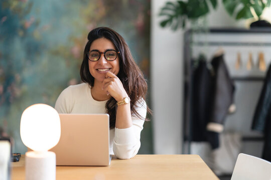A smiling woman with glasses sits at a desk with a laptop and a lamp, working in a modern office space.