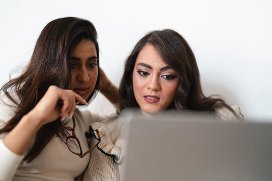 Two women are looking at a laptop screen together, one holding glasses and appearing thoughtful.