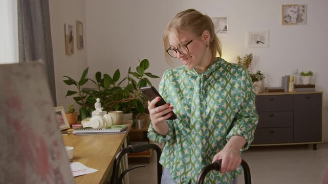 Tilt up view of joyful Caucasian young woman wearing blouse and jeans smiling while reading message on smartphone while standing with walker at home