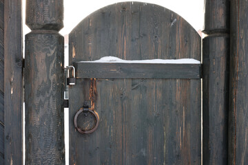 the wooden door is dark in color with metal elements. The hinges, deadbolt and ring are visible. There is some snow on the door