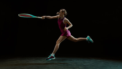 Athletic woman playing tennis in mid-air movement on dark stage. Concept of explosive performance, professional sport training, dynamic action and brand communication.