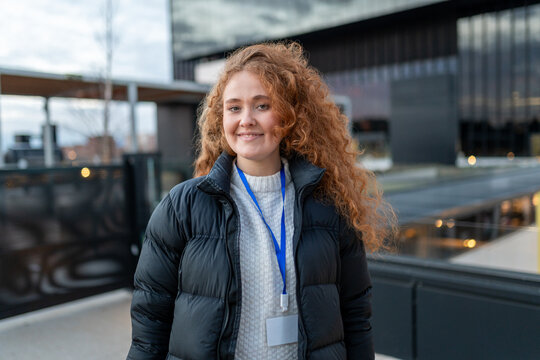 Young woman smiling, wearing a puffer jacket and blank id badge, standing outdoors in an urban setting