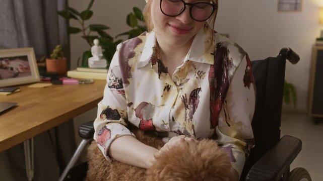 Tilt up view of smiling young woman with paraplegia petting small brown dog when sitting in wheelchair at home