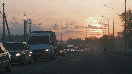 Evening traffic jam on an urban road at sunset. Cars with headlights on, busy city street, transportation congestion, rush hour, commuting and urban mobility concept.