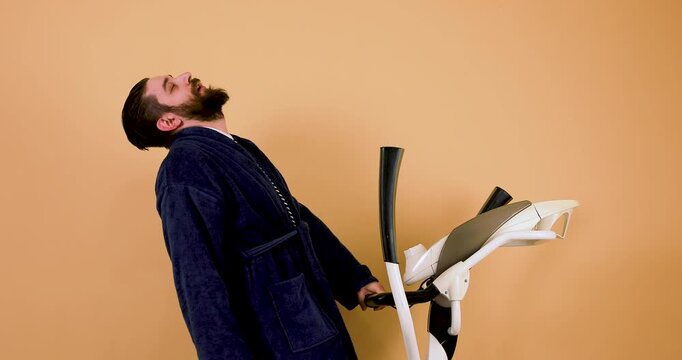 A bearded Caucasian man wearing a robe exercises on an elliptical trainer, emphasizing the concept of prioritizing fitness right after waking up. This 4K video represents discipline, healthy habits, m
