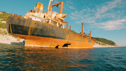 A rusty cargo ship with holes in its hull lies partially submerged in calm sea waters. The abandoned ship aground against a rocky hill. View from the water.