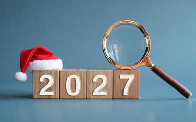 Wooden 2027 New Year Blocks with Santa Hat and Magnifying Glass on Blue Background for Future Forecast Concept