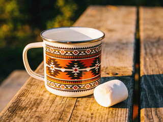 Enamel mug with tribal pattern and marshmallow on a wooden picnic table at sunset