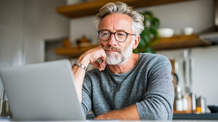 A senior man with glasses, looking at his laptop, deep in thought in his home kitchen.