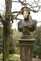 Weathered classical bust on a stone pedestal amid lush greenery in Stibbert Park, romantic English-style garden, Florence, Italy.