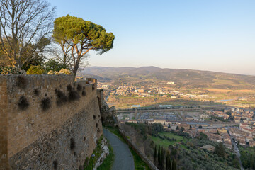 A view of a tranquil street in Orvieto, Italy, with traditional buildings and trees lining the path under the sunlight.