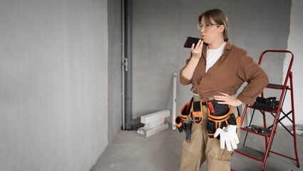 Female repairman using voice command on smartphone standing in empty room before renovation. profession, construction and building