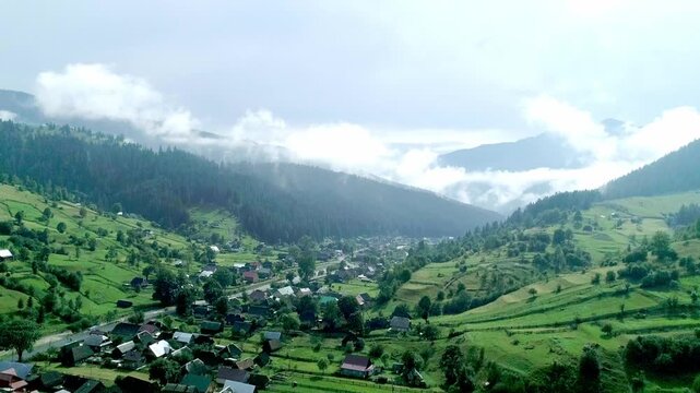 Aerial drone shot of serene mountain village nestled in lush green valley amid dense forests, winding river, patchwork fields and cozy red-roof homes. Vibrant summer countryside idyll for premium stoc