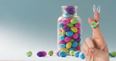 Image of fingers with easter bunny and easter egg over jar of easter eggs on blue background