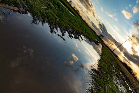 vista con orizzonte storto che mostra il cielo al tramonto che si riflette sull'acqua di una lunga pozzanghera, in un ambiente di campagna pianeggiante