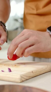 Vertical video: Reaching cook in orange tee picking blue knife at counter, slicing tomato for salad