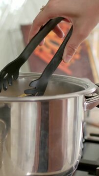 Vertical video: Lowering hand with black tongs placing fresh pasta into steel pot on stove, to cook
