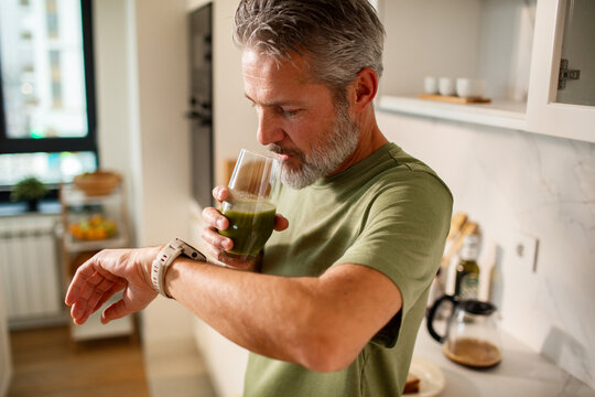Middle-aged man drinking green smoothie while checking smartwatch in home kitchen