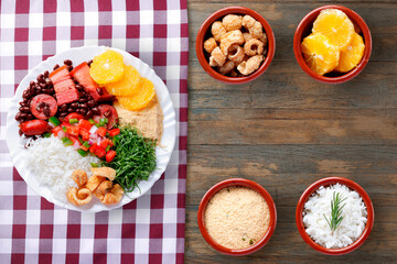 Complete Brazilian Feijoada with rice, collard greens, farofa, orange, and pork crackling served on a rustic wooden table with a checkered tablecloth